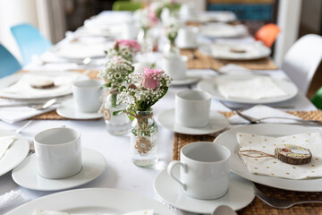 Table set for a wedding reception with white tablecloth, cups and plates with pink flowers in a glass. Decoration of table in dining room. 