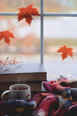 Cup of steaming hot tea wrapped in warm stack of books and maple leaves on window