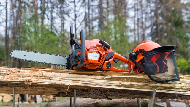 Close-up of portable chain saw and safety helmet on wood in metal sawhorse. Professional orange power chainsaw and protective hard hat with face shield of grid mesh on wooden log. Sawing machine tool.