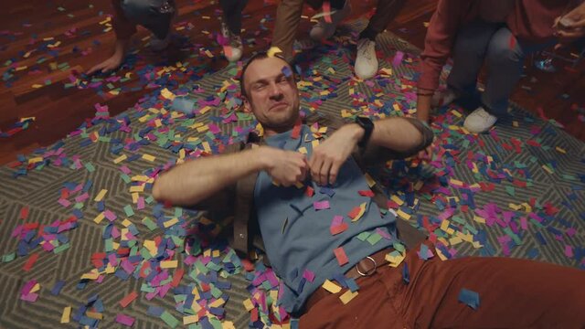 Slowmo of young cheerful man lying on floor throwing confetti to camera during friends home party