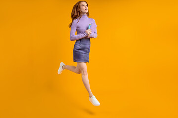 Fototapeta na wymiar Dreamy redhead lady at weekend jumping, hold notebooks in hands on way home, in air, wearing casual shirt and skirt isolated yellow orange background. Side view, copy space. Full-length portrait