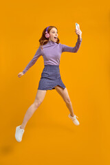 Fototapeta na wymiar Full length body size view of nice cheerful cheery woman in headhones jumping having fun using smartphone fast speed connection, listen to music, isolated on yellow background