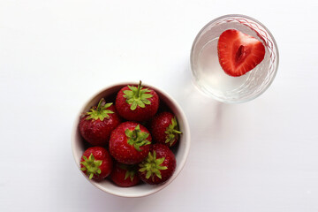Strawberry in a bowl. Glass of infused mineral water with half of strawberry on white table. Summer refreshing beverage with fresh berry. Top view.