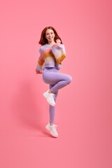 Attractive funny redhead lady jumping high raising hands first place winner wearing casual sweater, leggins isolated over bright pink color background. Beautiful lady rejoicing