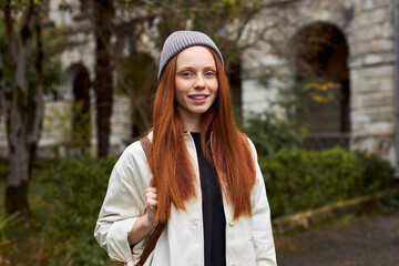 Close-up Portrait Of Young Woman With Natural Red Hair On Travel Trip, Outdoors, Posing At Camera, Smiling, In Coat And Hat. Old Building In The Background