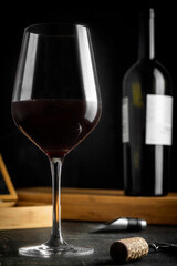 full wine glass on wooden table, with wine bottle behind, dark background and accessories on table.