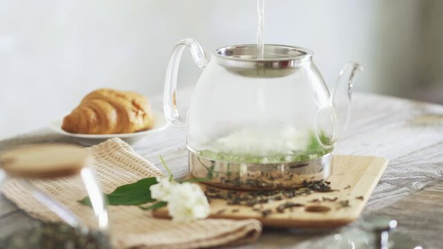 Pour hot water over jasmine flowers in teapot. Brew jasmine tea in transparent glass teapot. Pouring jasmine tea on a sunny morning. Cooking green tea croissant in the background. Hot drink concept.
