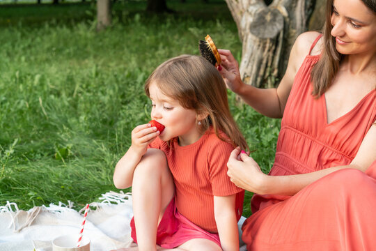 Happy smiling mother is combing her little daughter while sitting on plaid. Girl is eating ripe red strawberries.