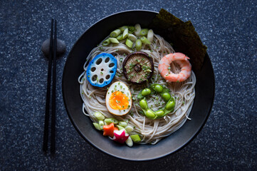 Bowl of Japanese buckwheat ramen noodle soup topped with colorful circles of blue lotus root, shiitake mushroom, shrimp, boiled egg, and edamame beans
