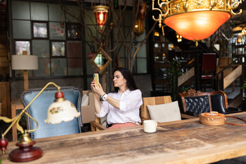 Happy young Caucasian millennial or gen z woman with long brunette hair streaming with smart phone in hands, shooting social media blog in modern cafe. Influencer using social networks indoor.