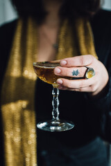 hand with mood ring and tattoos holding vintage champagne cocktail coupe glass and gold shimmering scarf - obrazy, fototapety, plakaty