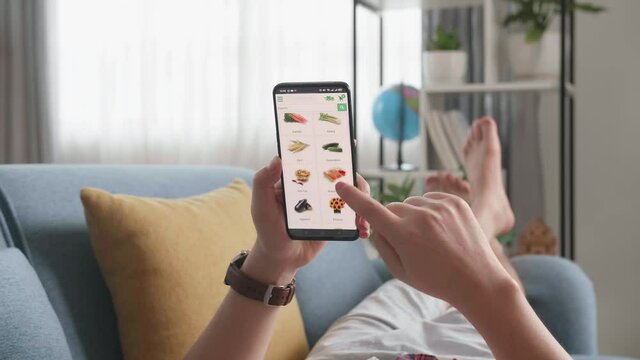 Man Lying On Couch Using Smartphone Buys In Internet Shop. Male Selects Vegetables In Grocery Online Store
