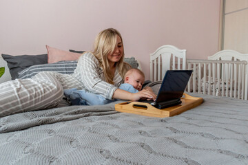 Young woman in comfortable home clothes work at laptop with a baby lying on bed in bedroom.