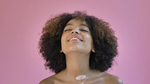 Relaxed black young woman with short curly hair massages neck with special jade massager roller against pink background closeup