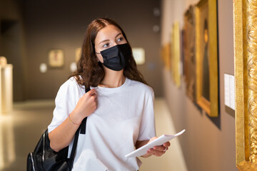 Portrait of a focused girl visitor wearing a protective mask, looking at paintings in the museum hall during the ..pandemic