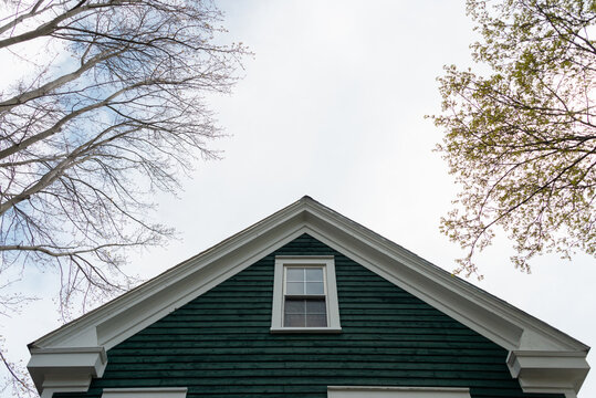A vintage blue wooden building with white trim, double hung window, dormer, a black shingled roof, and white wooden trim. The old home has multiple stories with a green tree and grey skies.