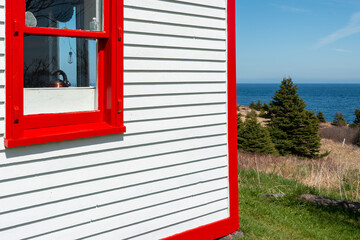 A white narrow wooden clapboard cape cod siding exterior corner wall of a house with bright red trim. There's a closed glass double hung window with red wood trim. A blue sky and ocean in the distance