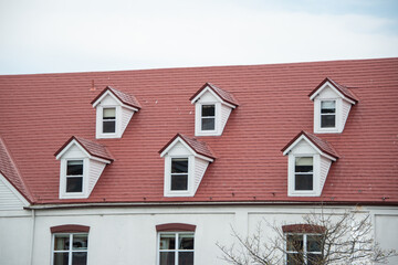 The corner of a red metal shingled roof with two raised dormers and three sunken small single windows in the exterior wall of a white stucco building. The vintage building has a clean design.
