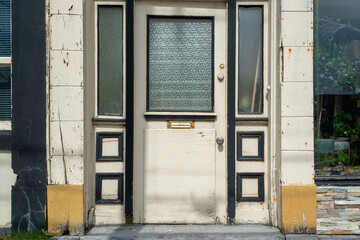 An old wooden cream color commercial door with half beveled glass on a building.  There are two decorative side windows and with wood panels. The trim is black with yellow and olive green color. 