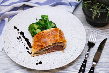 A slice of beef wellington with a portion of steamed broccoli dressed with the thick balsamic sauce on a white plate. Knife and fork on the side. Small cloth of napkin in a corner.