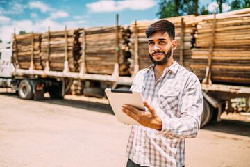 Portrait of Latin young man working with a digital tablet beside tree trunks on truck.