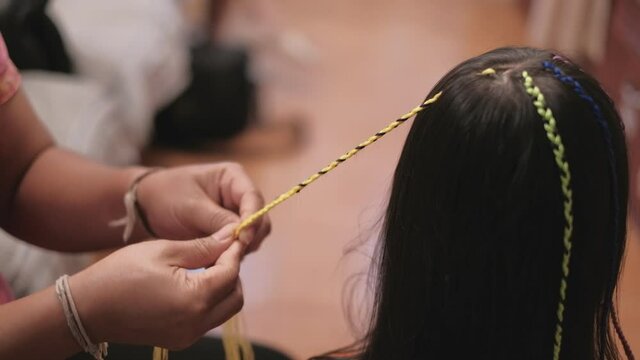 Close-up VDO: Cute long, black hair Asian girl is being braided in multi-colored threads by woman's hand in beauty salon. Child-like fashion, weaving pigtails with decoration yarn or knitting wool.