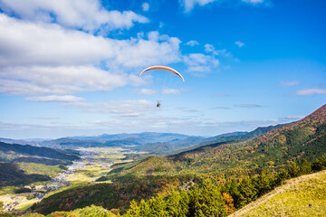 紅葉の山に囲まれた中で青空に向かって飛ぶパラグライダー。京都府与謝郡与謝野町