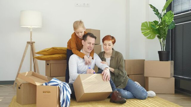 Young caucasian family moving to new place sitting holding marker pen writing carboard boxes, on the floor at home, enjoying time, on moving day. relocation concept