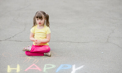 Little girl with syndrome down draws with chalk on the asphalt. Empty space for text