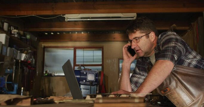 Focused caucasian male knife maker in workshop using laptop and smartphone