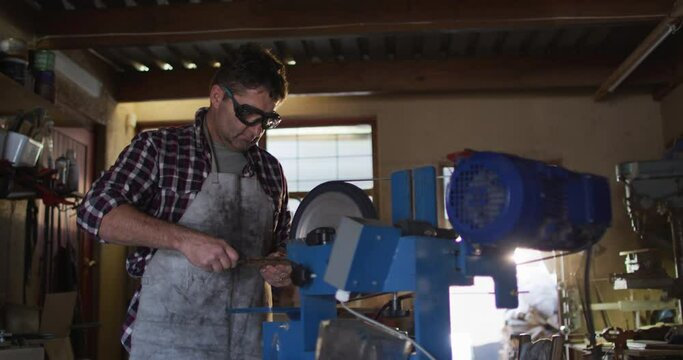 Caucasian male knife maker in workshop wearing glasses and using saw