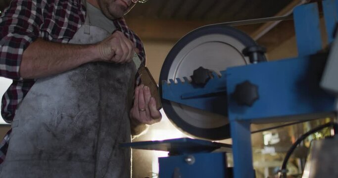 Caucasian male knife maker in workshop wearing glasses and using sander