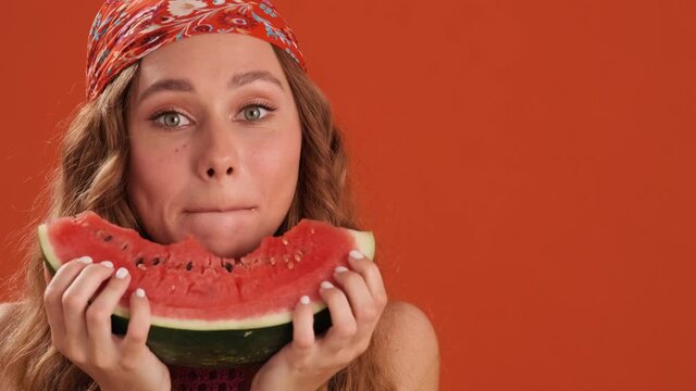 A close-up view of the curly-haired woman with bandana on her head biting a slice of watermelon in the orange studio
