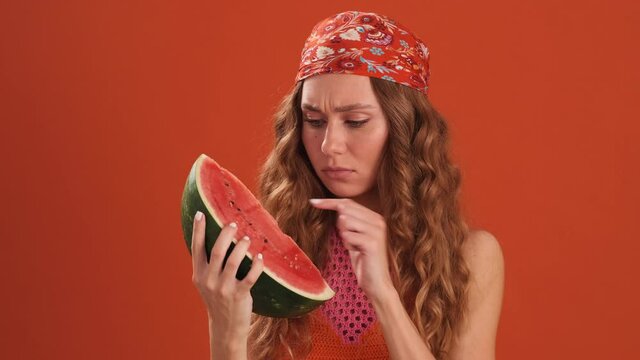 A laughing curly-haired woman with bandana on her head talking on a slice of watermelon like on the phone in the orange studio