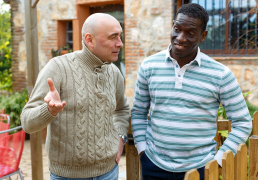 Neighbor conversation. Two smiling men breezily chatting near wooden wicket of rural house..