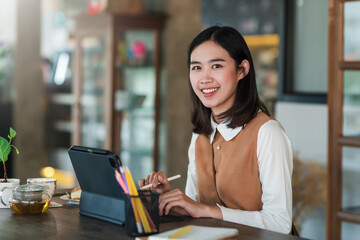 Young Asian woman working at a coffee shop holding a pen using a tablet. Looking at the camera.