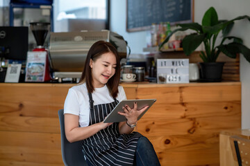 Small business owner Asian woman sitting in a chair in front of the counter happy wearing an apron using a tablet at a café.