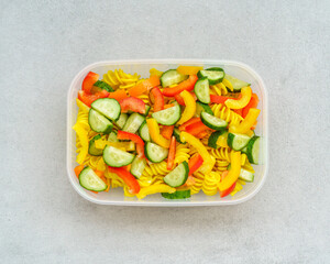 Top view on lunch box with vegetarian healthy vegetable cold pasta salad with cucumber and bell peppers isolated in grey background. Vegan recipes concept