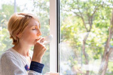 Fototapeta premium A woman drinking tea at the picture window with her eyes closed with pleasure