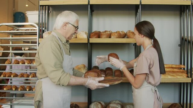 Slowmo dolly-out shot of cheerful senior baker holding tray with freshly baked bread and smiling while young female shopkeeper in apron and gloves putting loaves on shelf