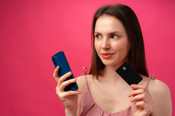 Smiling young woman holding smartphone and credit card against pink backgorund