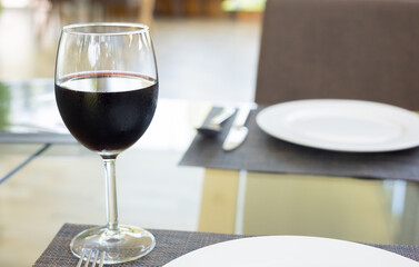 Selective focus and blurred background with copy space of glass of red wine with tableware clean set (white plate, fork and spoon) with prepared for food of lunch meal in luxury formal restaurant.
