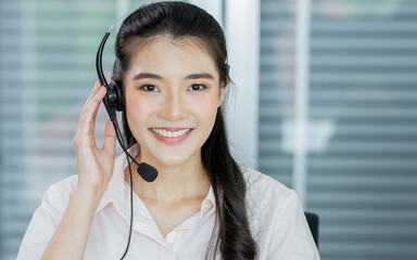 Portrait Asian beautiful attractive female call center cheerful smiling with headset, monitoring in office or agency at computer screen, giving service to customers, looking at camera with copy space.