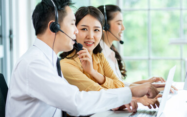 Selective focus Asian beautiful attractive female call center cheerful smiling with headsets, sitting, talking and consulting colleagues in office or agency at computer, giving service to customers.