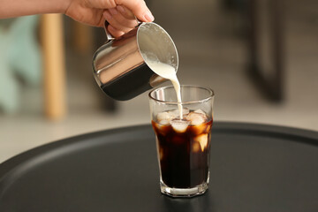 Woman pouring milk into glass with iced coffee on table