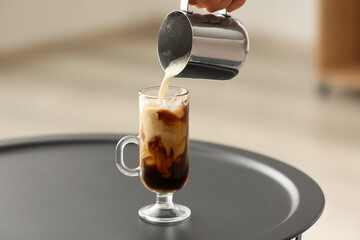 Woman pouring milk into glass with iced coffee on table, closeup
