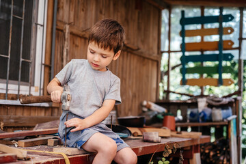 Fototapeta na wymiar Little boy hammers nails with a hammer in a wooden board in the carpenter's workshop