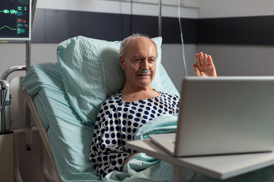 Portrait of senior man greeting family waving at laptop camera laying in hospital bed, after illness diagnosis, breathing with oxygen tube. Modern eqipment monitoring pacient heart rate during
