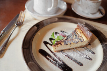 cheesecake on a plate in a restaurant or cafe on the table. the dessert is decorated with mint leaves, strawberries, chocolate syrup and powdered sugar. a fork, a teacup in the background. selective 