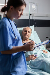 Medical nurse in scrubs taking notest on cliboard after consultation of sick unwell senior man. Patient with iv drip attached breathing with help from oxygen mask.